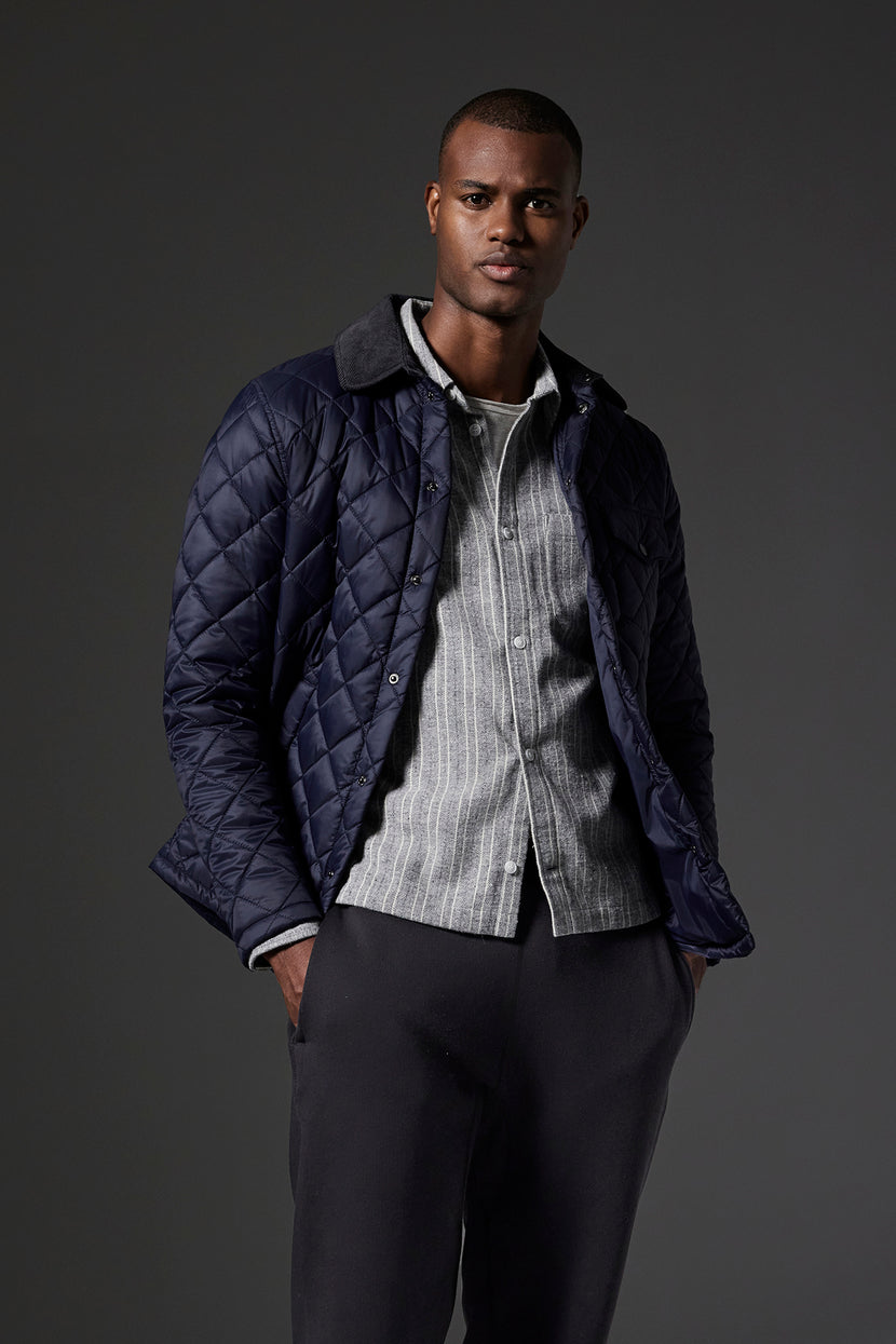 A man wearing the LOU JACKET, a dark quilted puffer jacket with a corduroy collar by Velvet by Graham & Spencer, over a light-colored shirt and dark pants stands against a dark background, with one hand in his pocket.