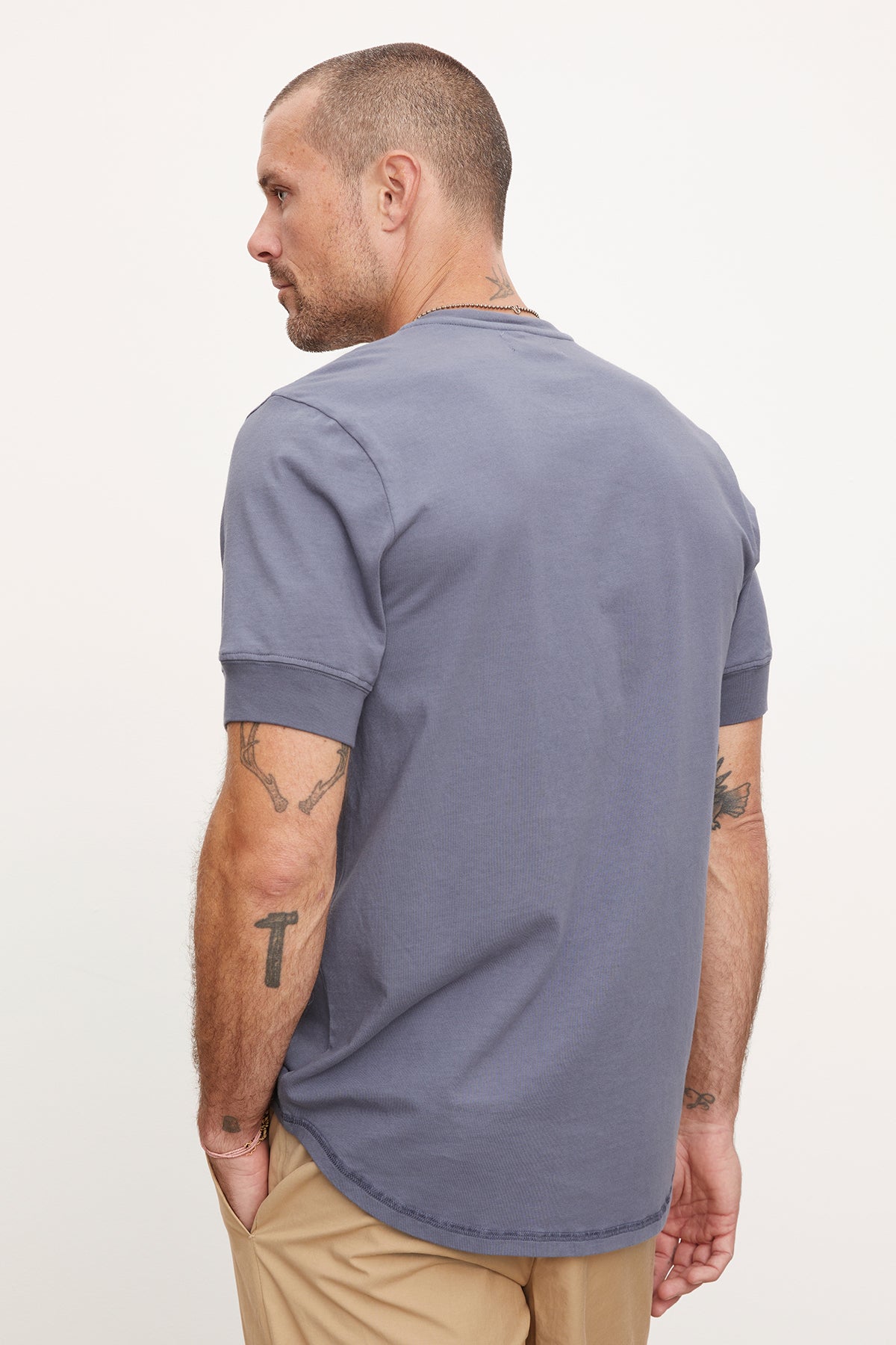 A man in a Velvet by Graham & Spencer DEON Henley t-shirt with scooped hem and beige pants, viewed from the side as he looks to the left, displaying tattoos on his arms.-36918612000961