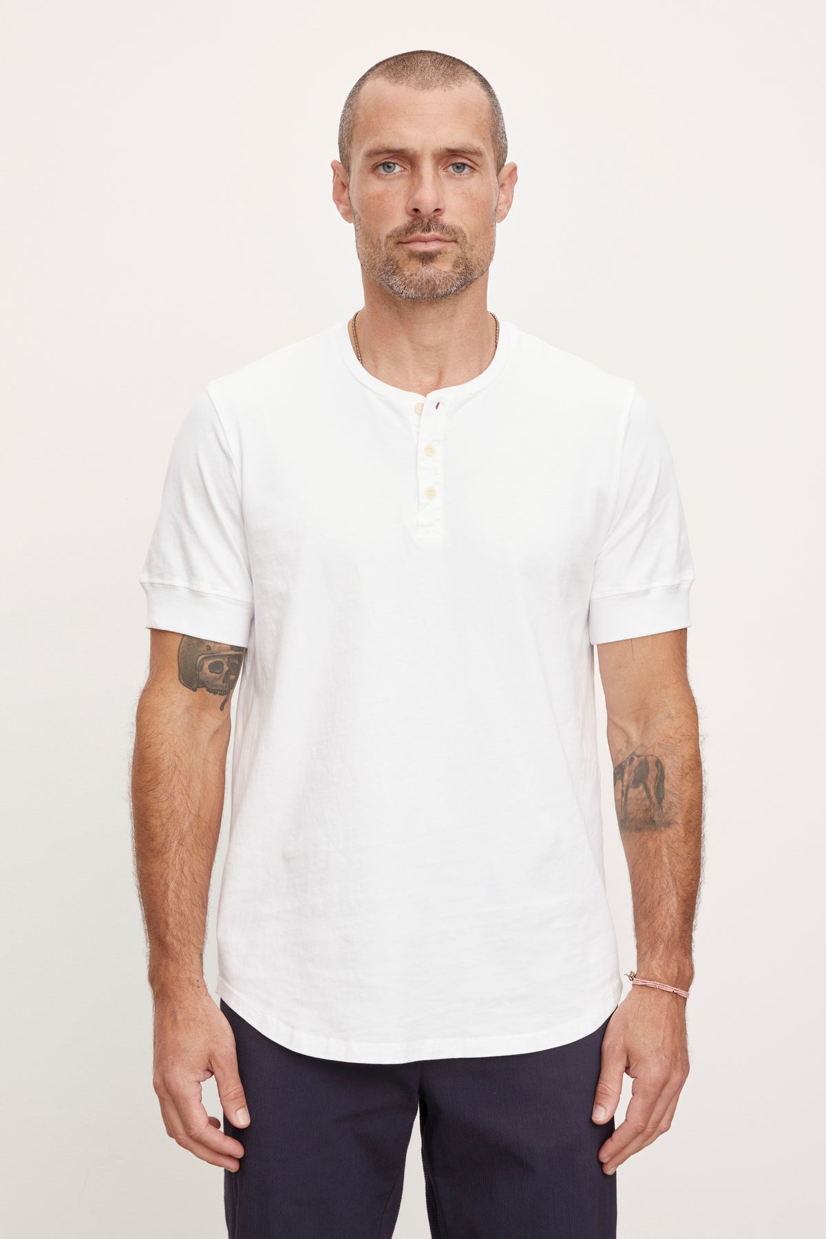 A man with a short haircut and tattoos on his arms stands against a plain background, wearing a white cotton knit DEON HENLEY by Velvet by Graham & Spencer with scooped hem details and dark pants.-36753582948545
