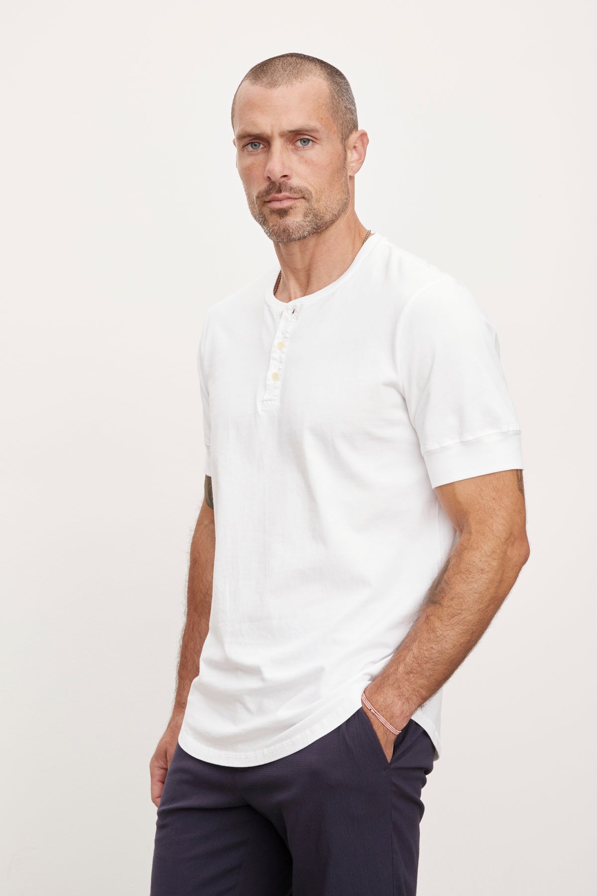 A man with a beard in a white DEON HENLEY shirt by Velvet by Graham & Spencer and navy trousers stands with hands in pockets, looking directly at the camera.-36753582850241