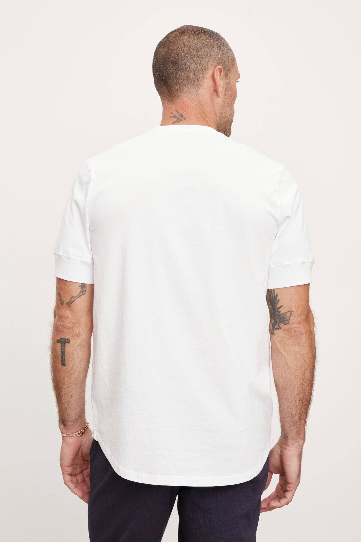 A man viewed from behind wearing a plain white short-sleeved Velvet by Graham & Spencer DEON HENLEY t-shirt and navy trousers, with visible tattoos on both arms.-36753582883009