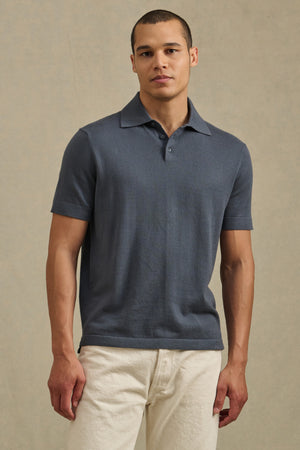 A man faces forward against a plain background, wearing the Velvet by Graham & Spencer CLYDE SWEATER POLO—a resort-ready, short-sleeve lightweight blue sweater polo—paired with light-colored pants.