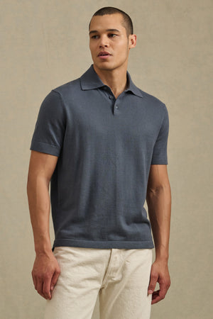 A man wears the CLYDE SWEATER POLO by Velvet by Graham & Spencer in blue with white pants, standing against a plain beige background for a relaxed, resort-ready look.
