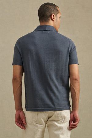 A man faces away from the camera, wearing the Velvet by Graham & Spencer CLYDE SWEATER POLO in dark gray with light pants, set against a plain background.