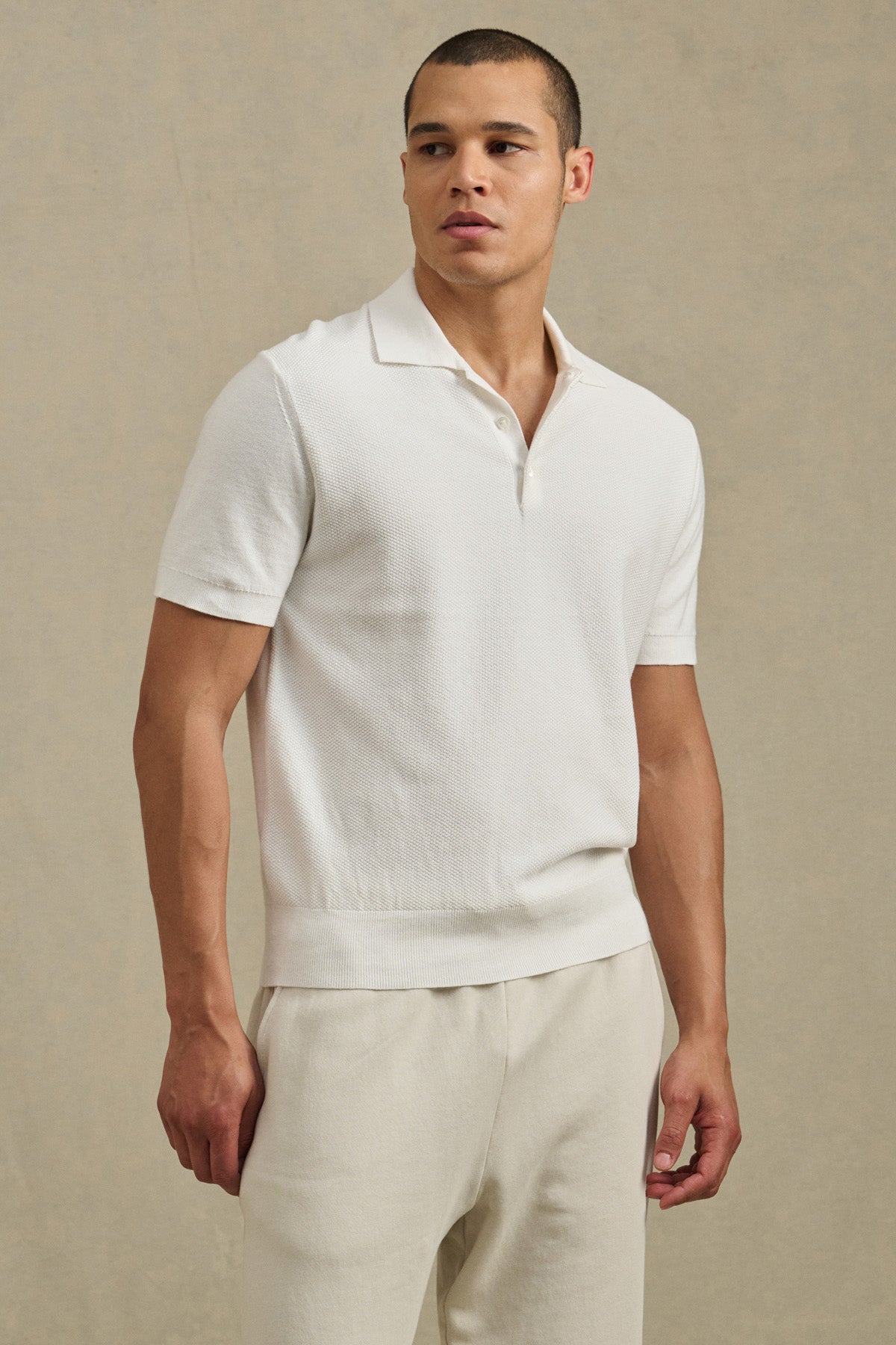 A man showcases the DILLON SWEATER POLO by Velvet by Graham & Spencer—its white textured design pairs effortlessly with light pants against a beige backdrop, creating a chic warm-weather look.-41428107526337