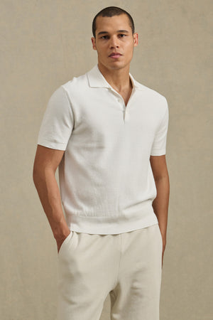 A man stands against a neutral background wearing the DILLON SWEATER POLO by Velvet by Graham & Spencer—an ideal warm-weather layer—in white, paired with off-white pants and his hands in his pockets.