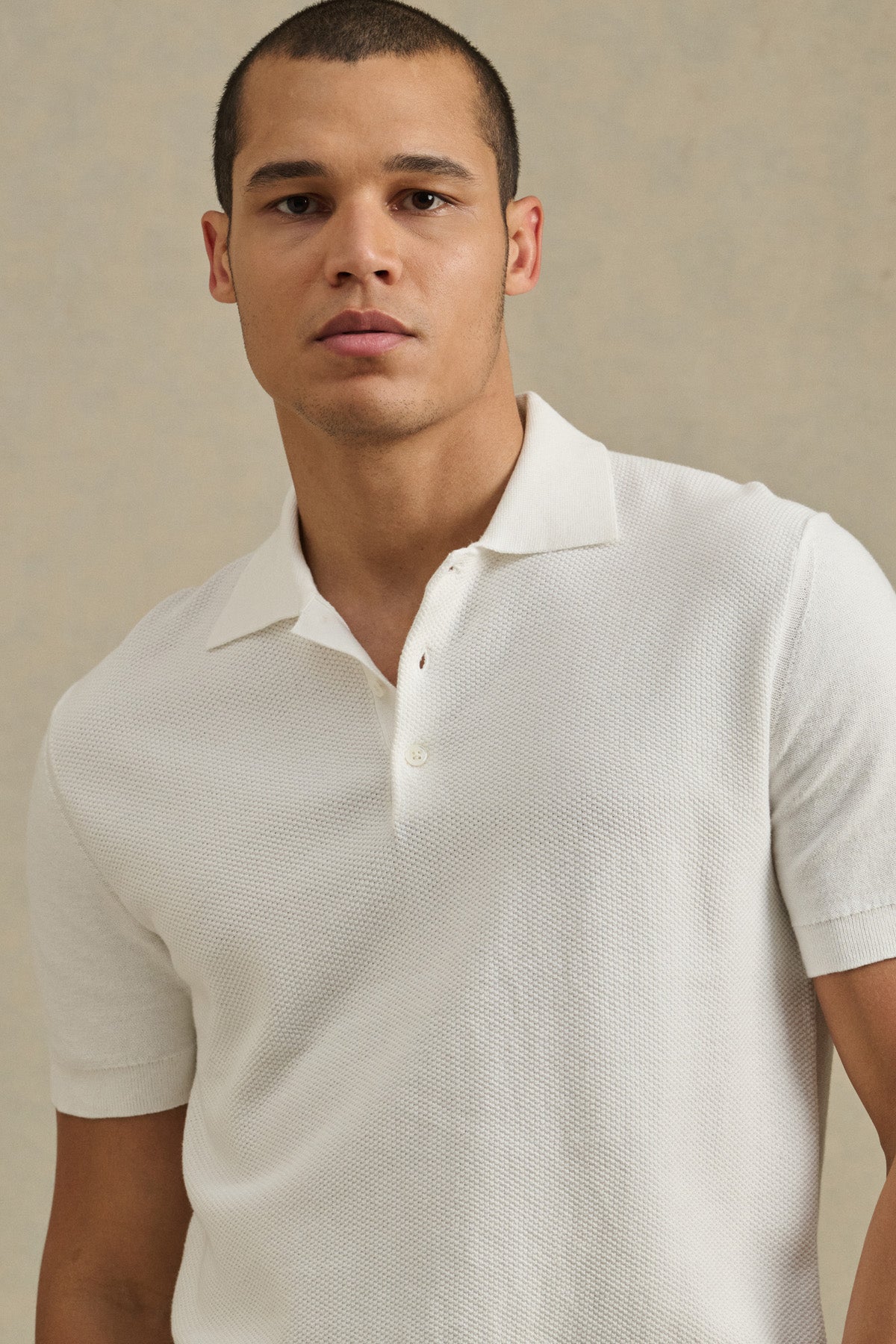 A man wears the DILLON SWEATER POLO by Velvet by Graham & Spencer, standing against a beige background—a stylish, textured layer ideal for warm weather.-41428107591873