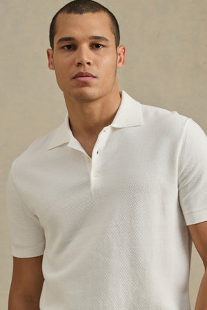 A man wears the DILLON SWEATER POLO by Velvet by Graham & Spencer, standing against a beige background—a stylish, textured layer ideal for warm weather.