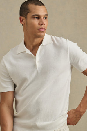 A man models the DILLON SWEATER POLO by Velvet by Graham & Spencer—a short-sleeved, textured piece—against a beige background, looking left with one hand on his hip. Perfect as a warm-weather layer.