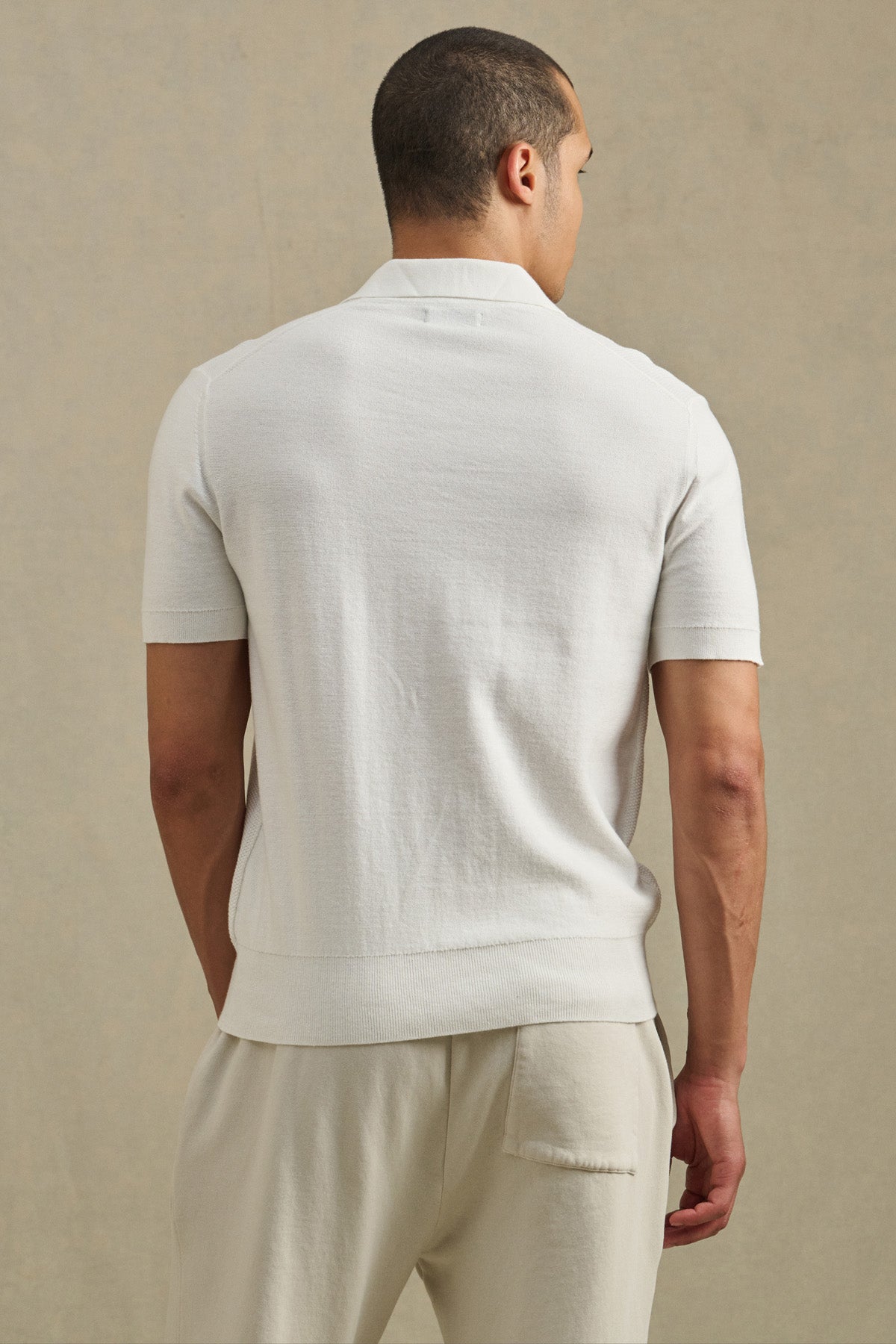 A man stands facing away, wearing the DILLON SWEATER POLO by Velvet by Graham & Spencer with matching light pants—a refined warm-weather layer set against a neutral background.-41428107690177