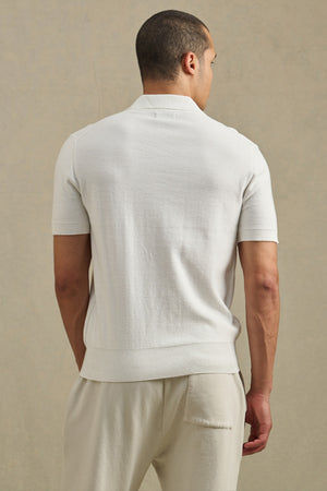 A man stands facing away, wearing the DILLON SWEATER POLO by Velvet by Graham & Spencer with matching light pants—a refined warm-weather layer set against a neutral background.