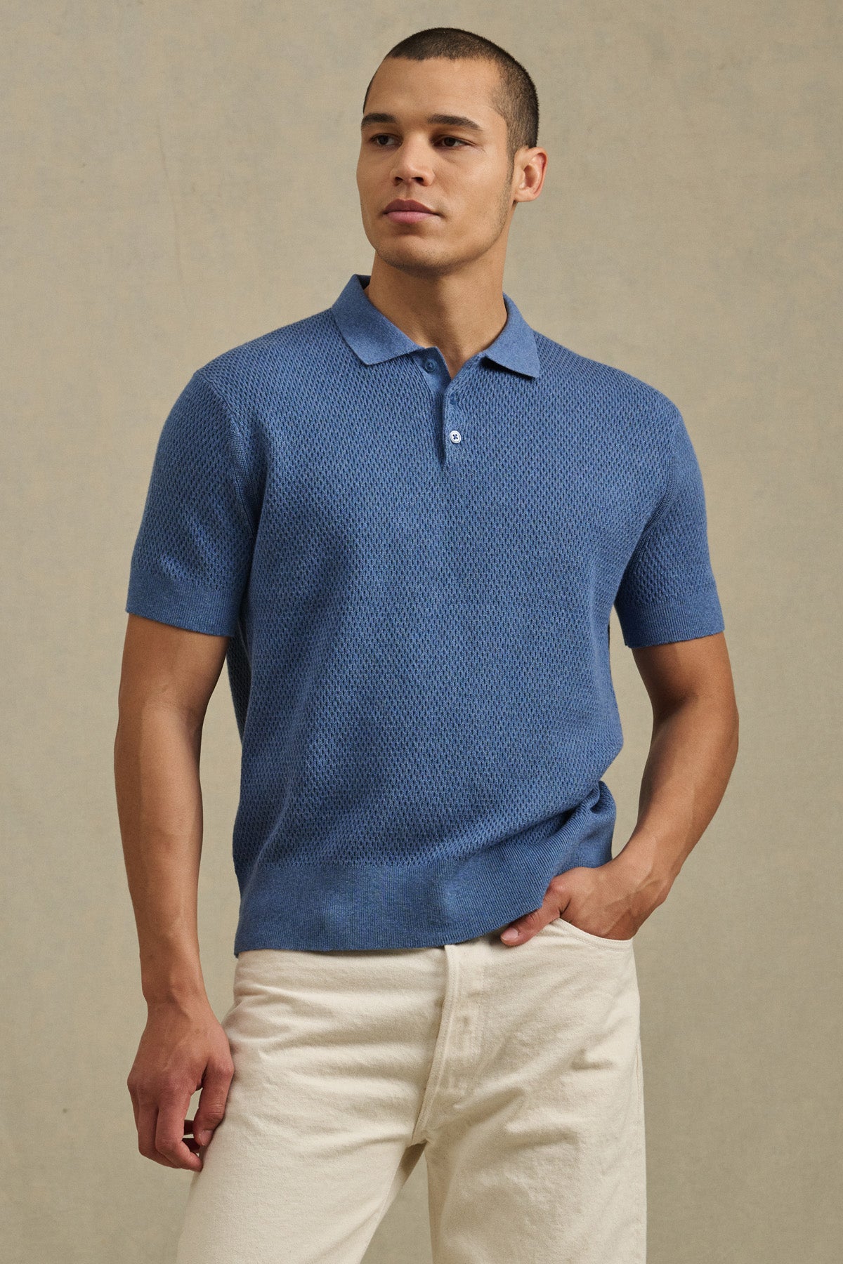 A man stands against a plain beige background wearing the LOWELL SWEATER POLO by Velvet by Graham & Spencer in textured blue and off-white pants, showcasing this versatile wardrobe staple.-41145319391425