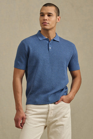 A man stands against a plain beige background wearing the LOWELL SWEATER POLO by Velvet by Graham & Spencer in textured blue and off-white pants, showcasing this versatile wardrobe staple.