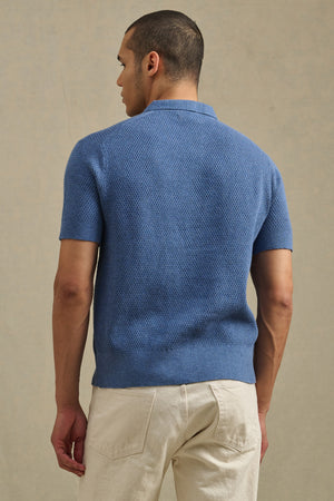 A man faces away, wearing the LOWELL SWEATER POLO by Velvet by Graham & Spencer—a textured blue cotton-cashmere piece—paired with light pants, set against a neutral background.