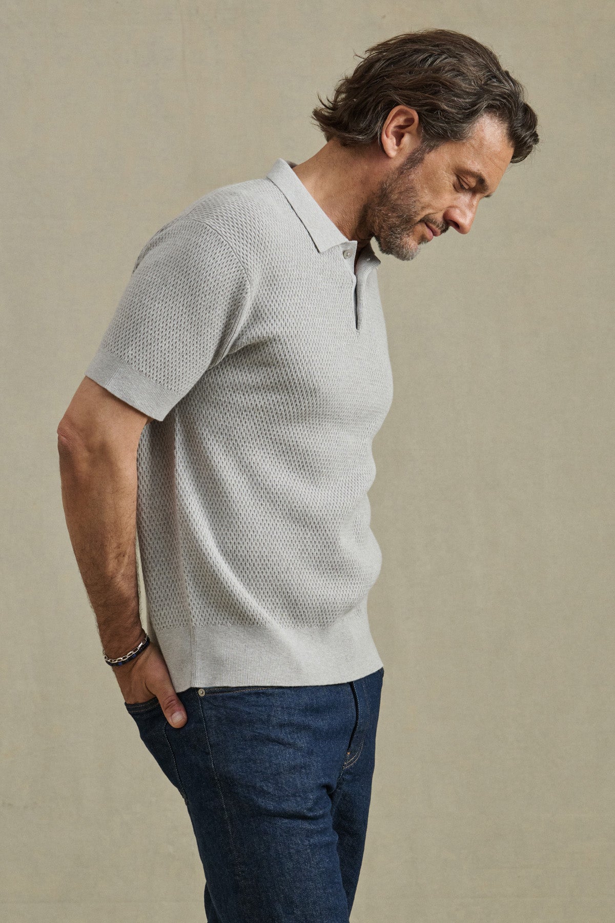 A man wears the LOWELL SWEATER POLO by Velvet by Graham & Spencer in light gray with breathable mesh, paired with dark blue jeans, standing in profile with hands in pockets against a plain beige background.