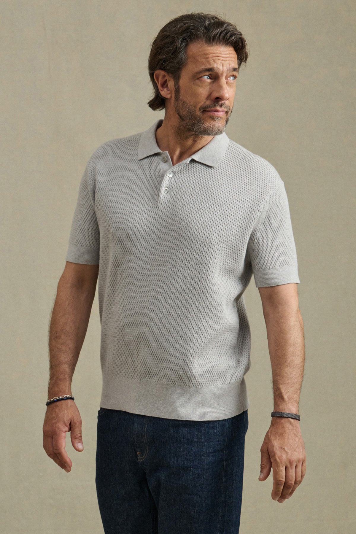 Man wearing the LOWELL SWEATER POLO by Velvet by Graham & Spencer in light gray with dark blue jeans, standing against a plain beige background and looking to his left.