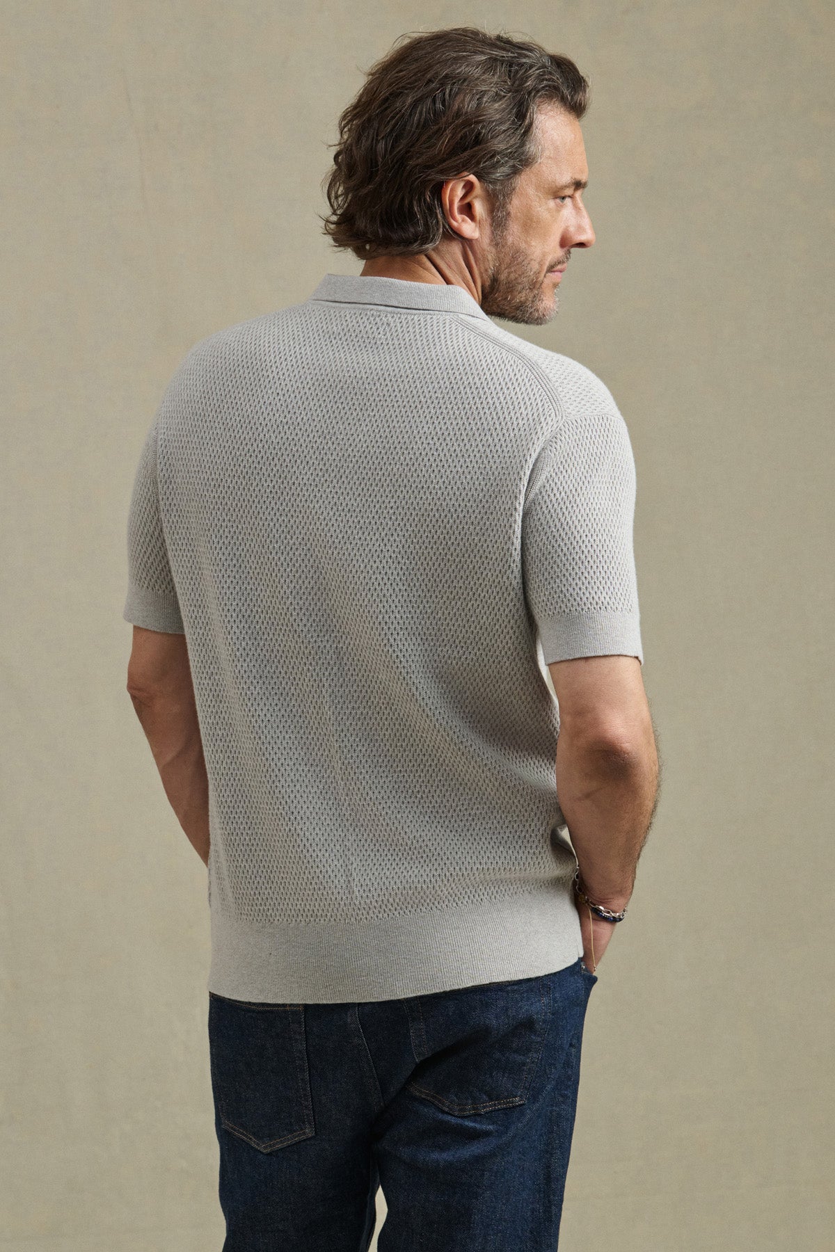 A man stands with his back to the camera in a light gray LOWELL SWEATER POLO by Velvet by Graham & Spencer and dark blue jeans, set against a plain beige background.