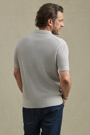 A man stands with his back to the camera in a light gray LOWELL SWEATER POLO by Velvet by Graham & Spencer and dark blue jeans, set against a plain beige background.