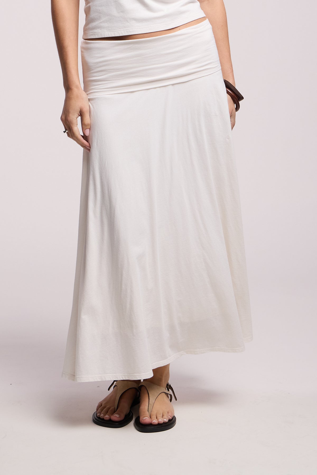 A person wears the LUELLA SKIRT by Velvet by Graham & Spencer—a long white A-line silhouette—paired with a white top, brown bracelet, and black sandals, standing against a plain light background.