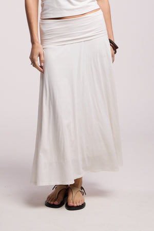 A person wears the LUELLA SKIRT by Velvet by Graham & Spencer—a long white A-line silhouette—paired with a white top, brown bracelet, and black sandals, standing against a plain light background.