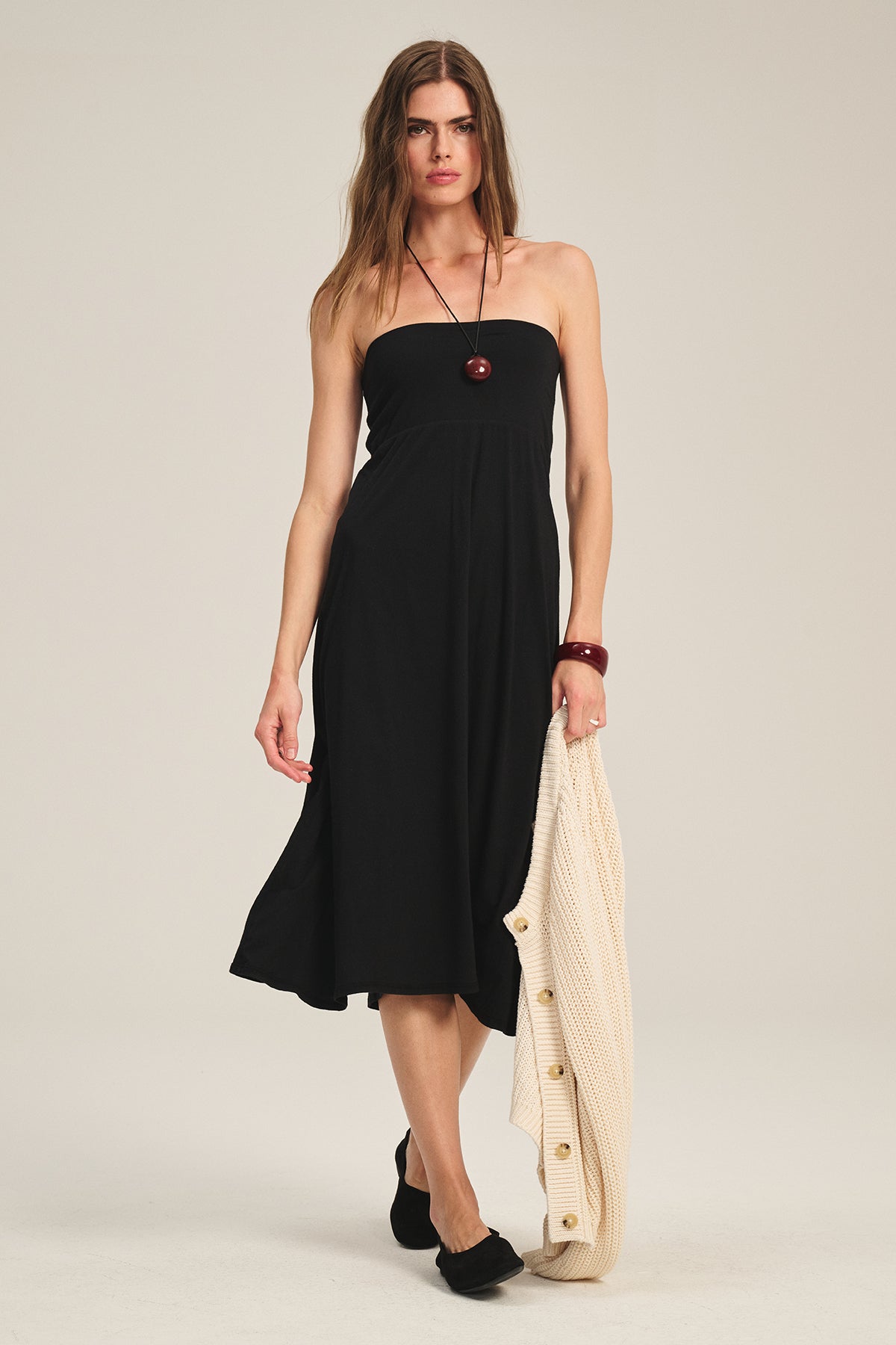 A woman wears the LUELLA SKIRT by Velvet by Graham & Spencer, styled with black flats and holds a cream buttoned cardigan, standing against a plain background.-39600485761217