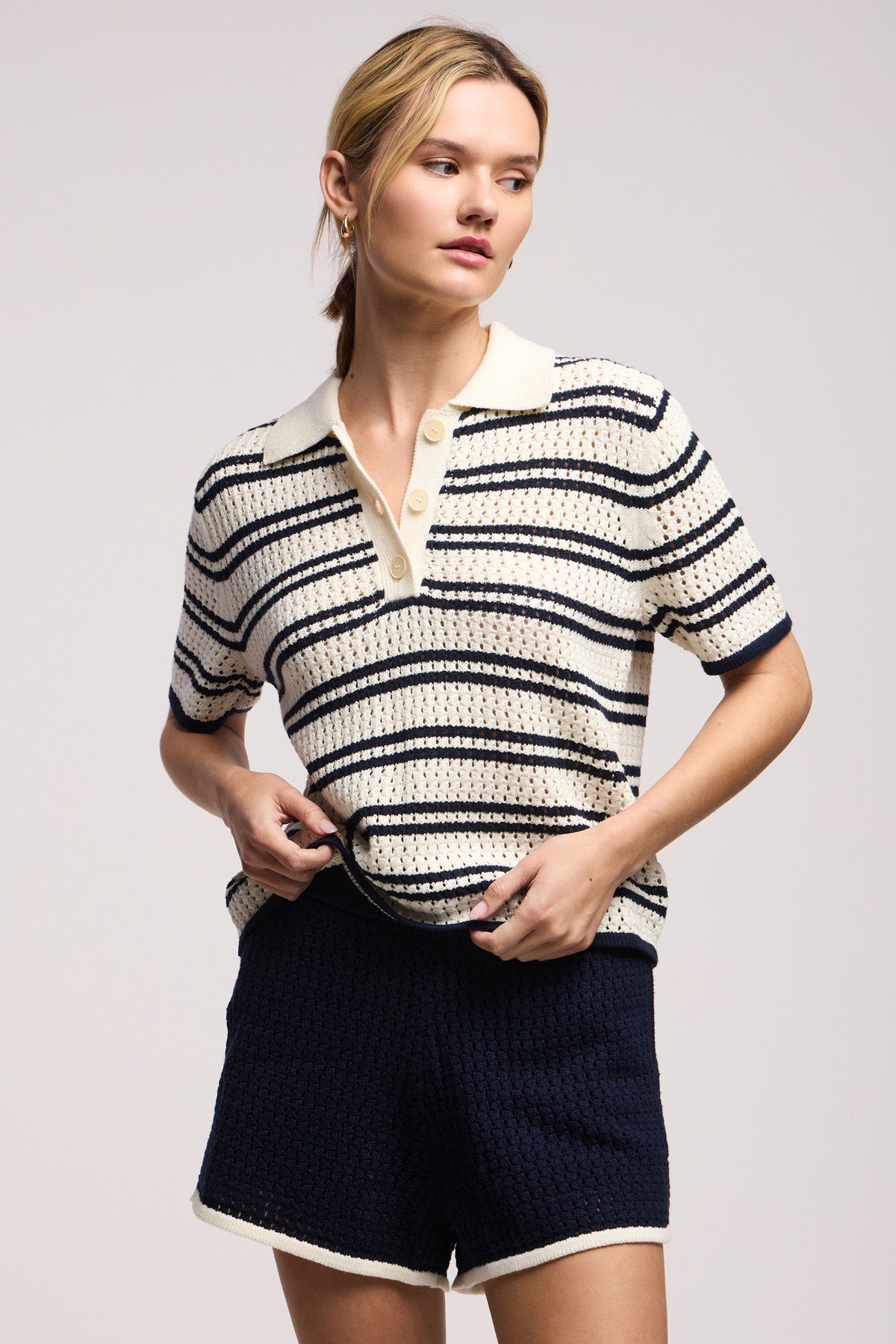 A woman wears the LUMAE SWEATER POLO by Velvet by Graham & Spencer—a short-sleeve, collared, button-up striped crochet top with contrast piping—paired with matching dark shorts with light trim, standing against a plain background.-41371964309697