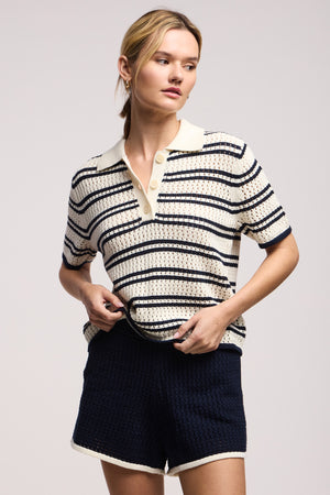 A woman wears the LUMAE SWEATER POLO by Velvet by Graham & Spencer—a short-sleeve, collared, button-up striped crochet top with contrast piping—paired with matching dark shorts with light trim, standing against a plain background.