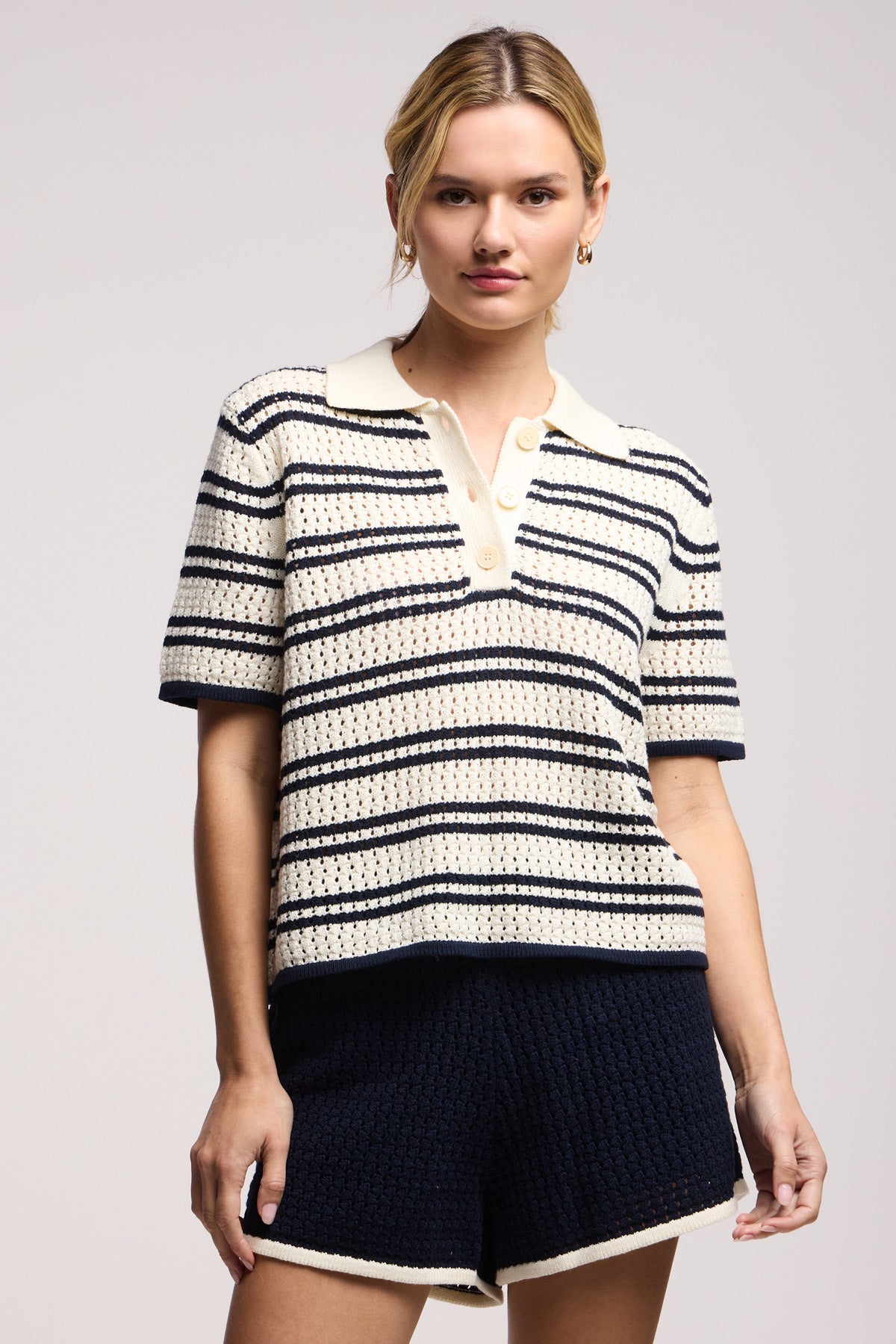 A woman wears the LUMAE SWEATER POLO by Velvet by Graham & Spencer, a sporty-chic knit top with horizontal navy and cream stripes and contrast piping, paired with high-waisted navy knit shorts against a plain background.-41371964342465