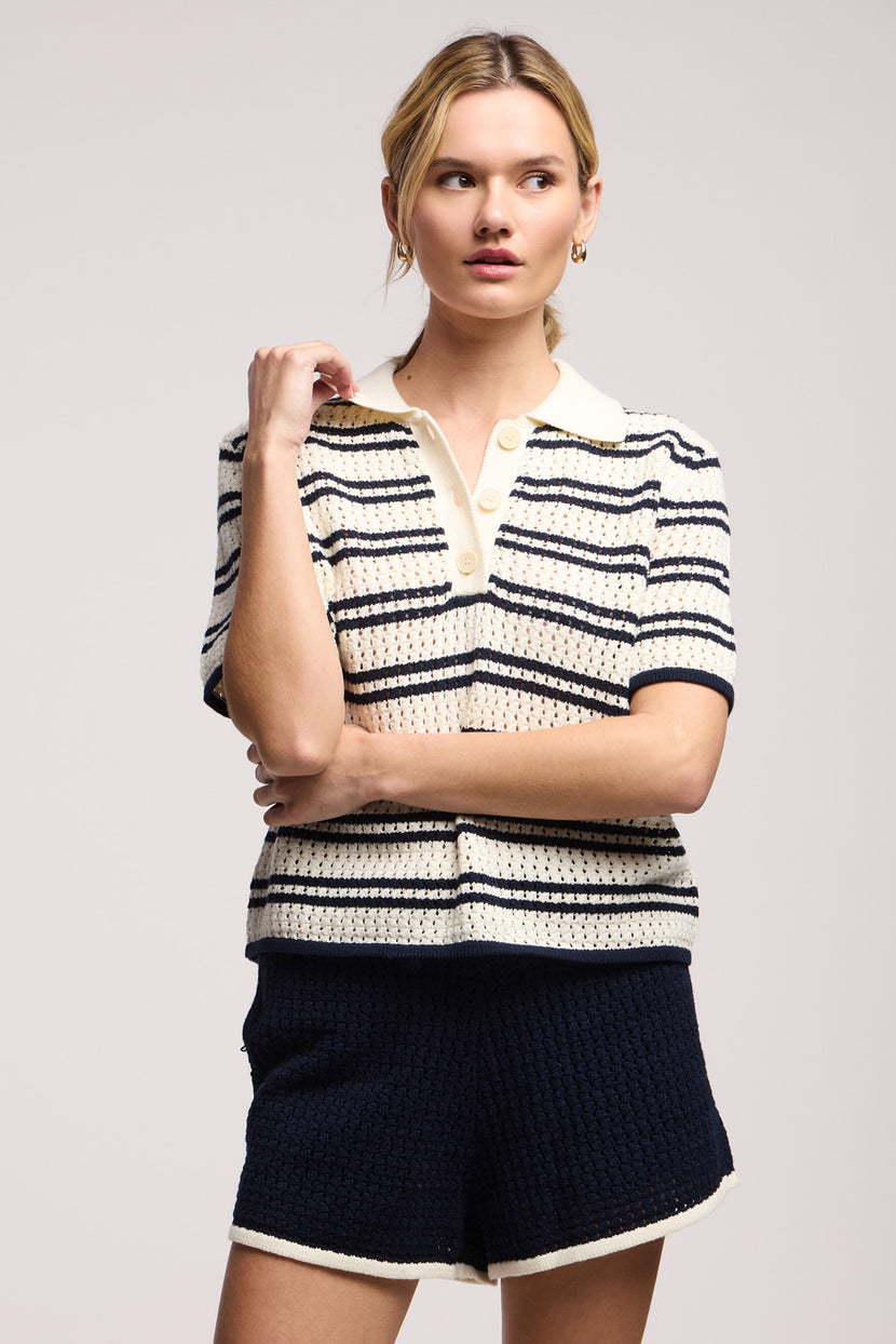 A woman stands with one hand on her chin, wearing the LUMAE SWEATER POLO by Velvet by Graham & Spencer—a sporty-chic knit top with contrast piping—paired with dark knit shorts featuring white trim.