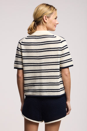A woman with blonde hair in a low ponytail stands sideways, wearing the Velvet by Graham & Spencer LUMAE SWEATER POLO in cream and navy stripes paired with navy textured shorts featuring cream trim.