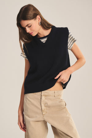 Wearing the LUMINA SWEATER VEST by Velvet by Graham & Spencer in navy over a striped tee and beige pants, a person glances downward and lifts the vest slightly to show the waistband.