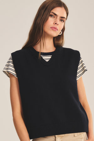 A woman wears the LUMINA SWEATER VEST by Velvet by Graham & Spencer in navy over a black-and-white striped short-sleeve top, creating a chic layered look against a neutral background.