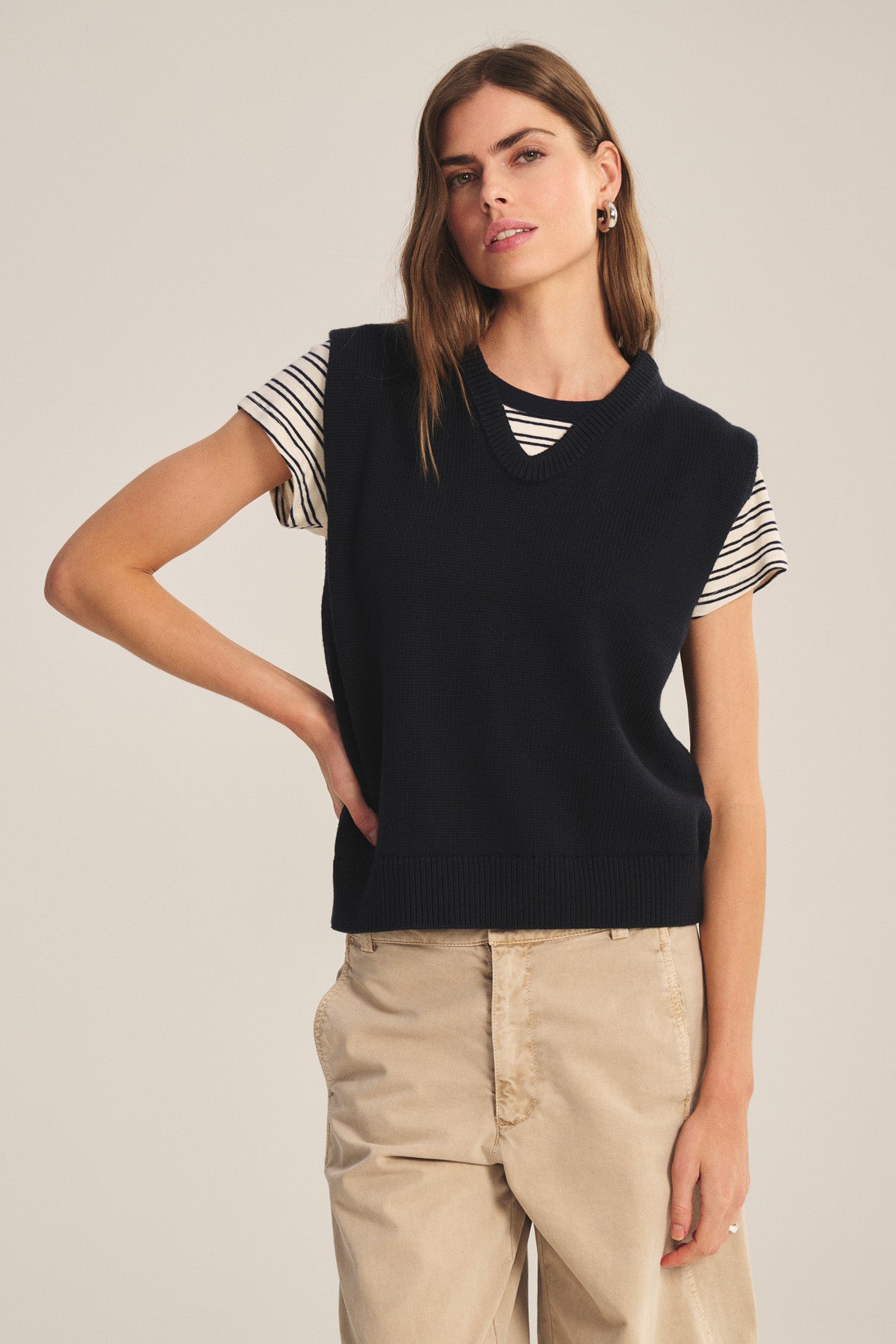A woman stands against a plain background, wearing the LUMINA SWEATER VEST by Velvet by Graham & Spencer layered over a striped short-sleeve shirt and paired with beige pants.-40909856047297