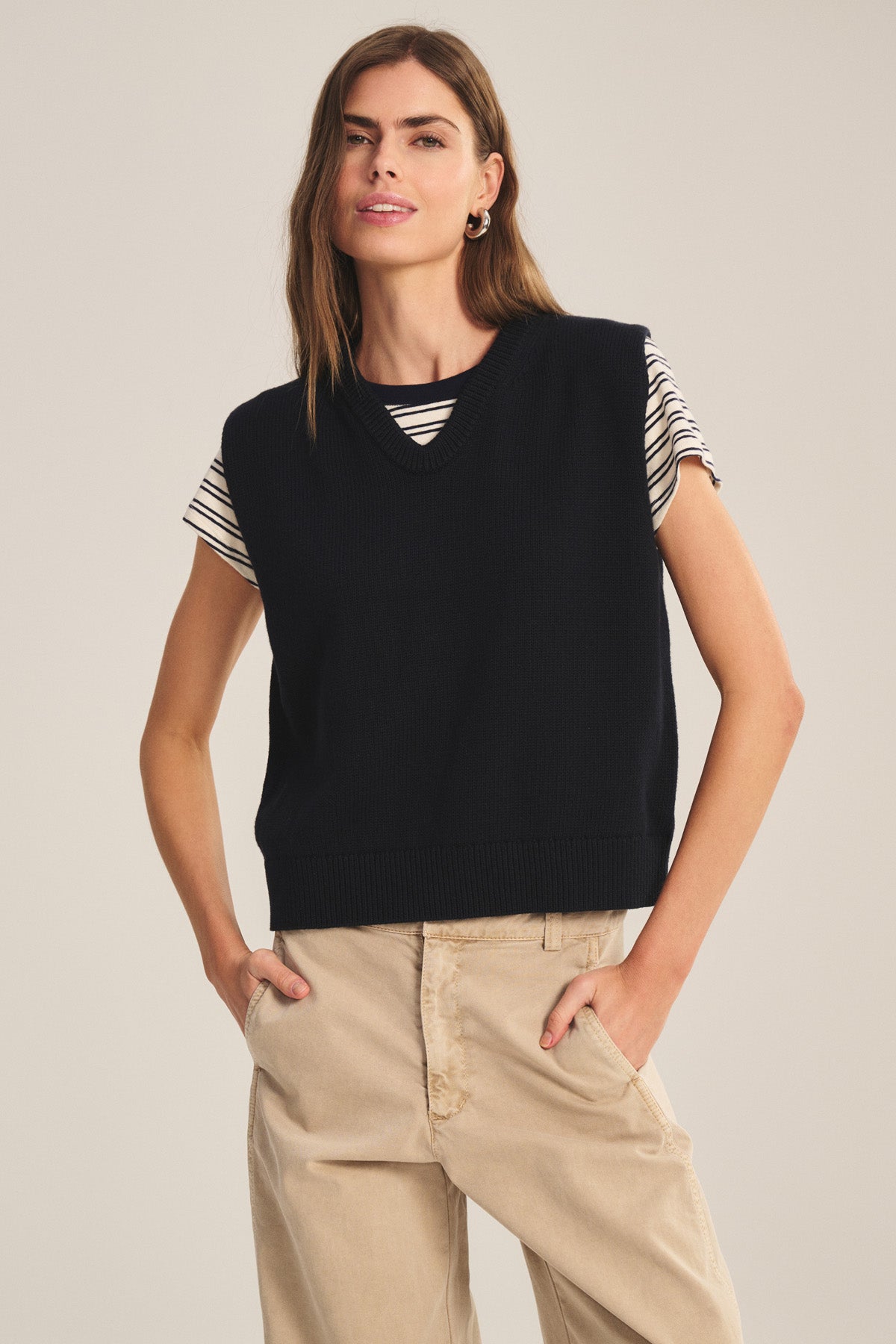 A woman stands with one hand in her pocket, wearing the LUMINA SWEATER VEST by Velvet by Graham & Spencer over a striped t-shirt and beige pants for a chic layered look.