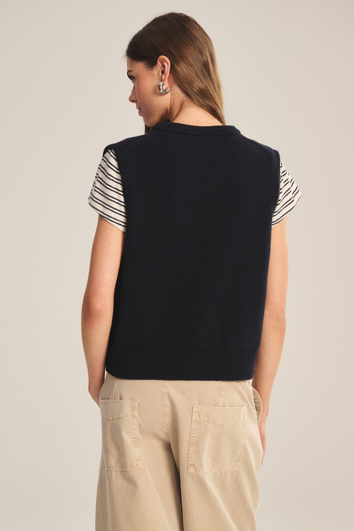 A woman faces away, wearing the LUMINA SWEATER VEST by Velvet by Graham & Spencer over a black and white striped tee and beige pants with large back pockets for a chic, layered look.-40909855981761