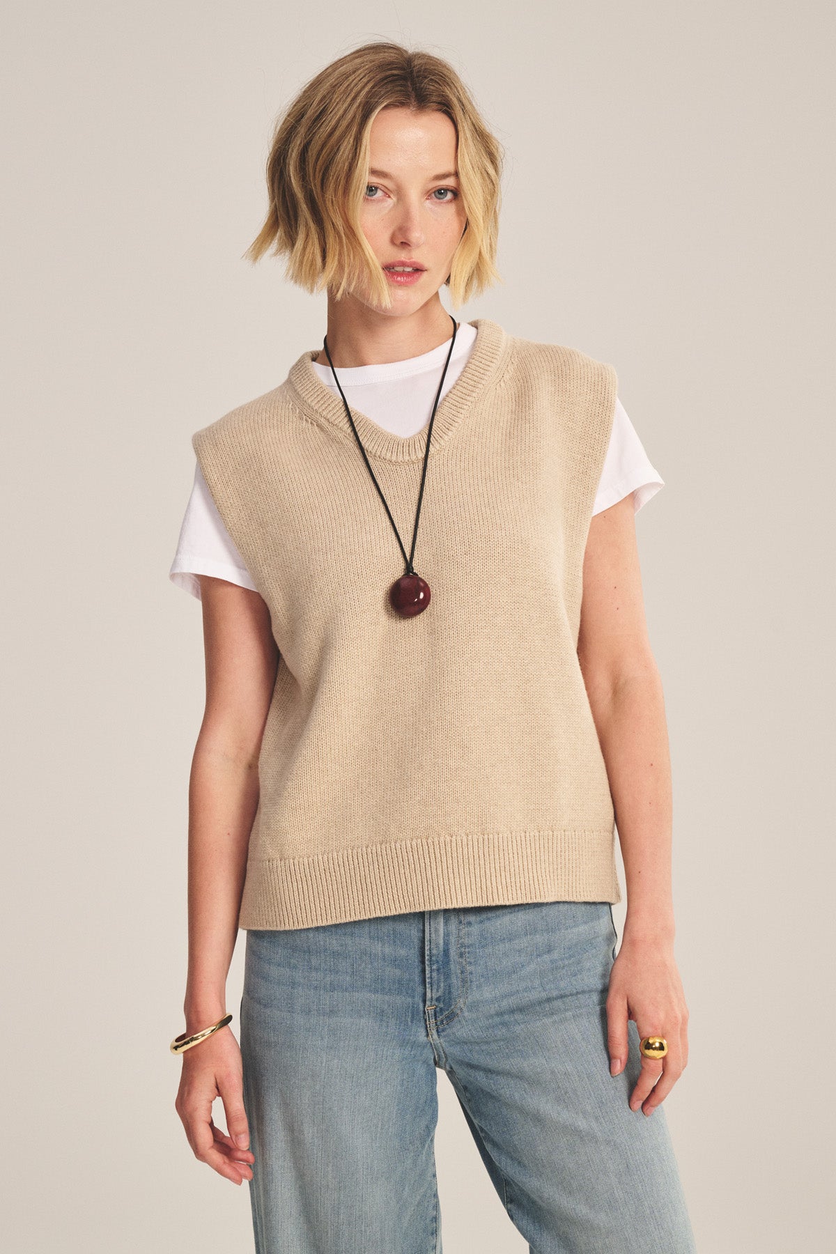 A person models the Velvet by Graham & Spencer LUMINA SWEATER VEST over a white tee with light blue jeans, a black round pendant necklace, and gold jewelry, creating a stylish layered look against a plain background.-40909855948993