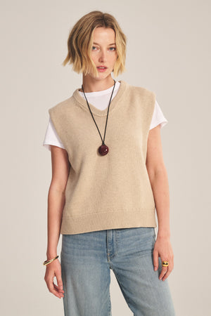 A person models the Velvet by Graham & Spencer LUMINA SWEATER VEST over a white tee with light blue jeans, a black round pendant necklace, and gold jewelry, creating a stylish layered look against a plain background.
