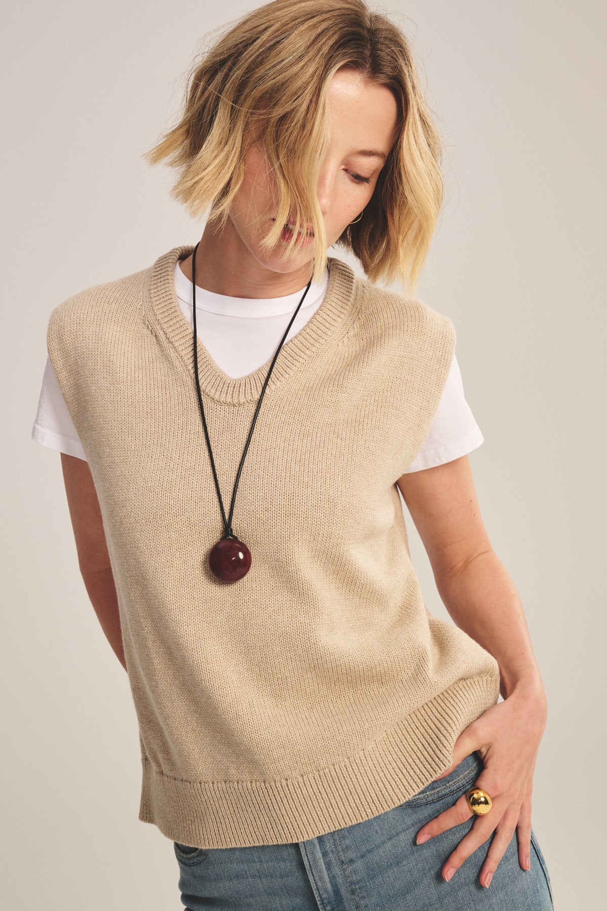 A person wears the LUMINA SWEATER VEST by Velvet by Graham & Spencer over a white t-shirt, paired with blue jeans, a large pendant necklace, and a gold ring against a plain background.-40909855916225