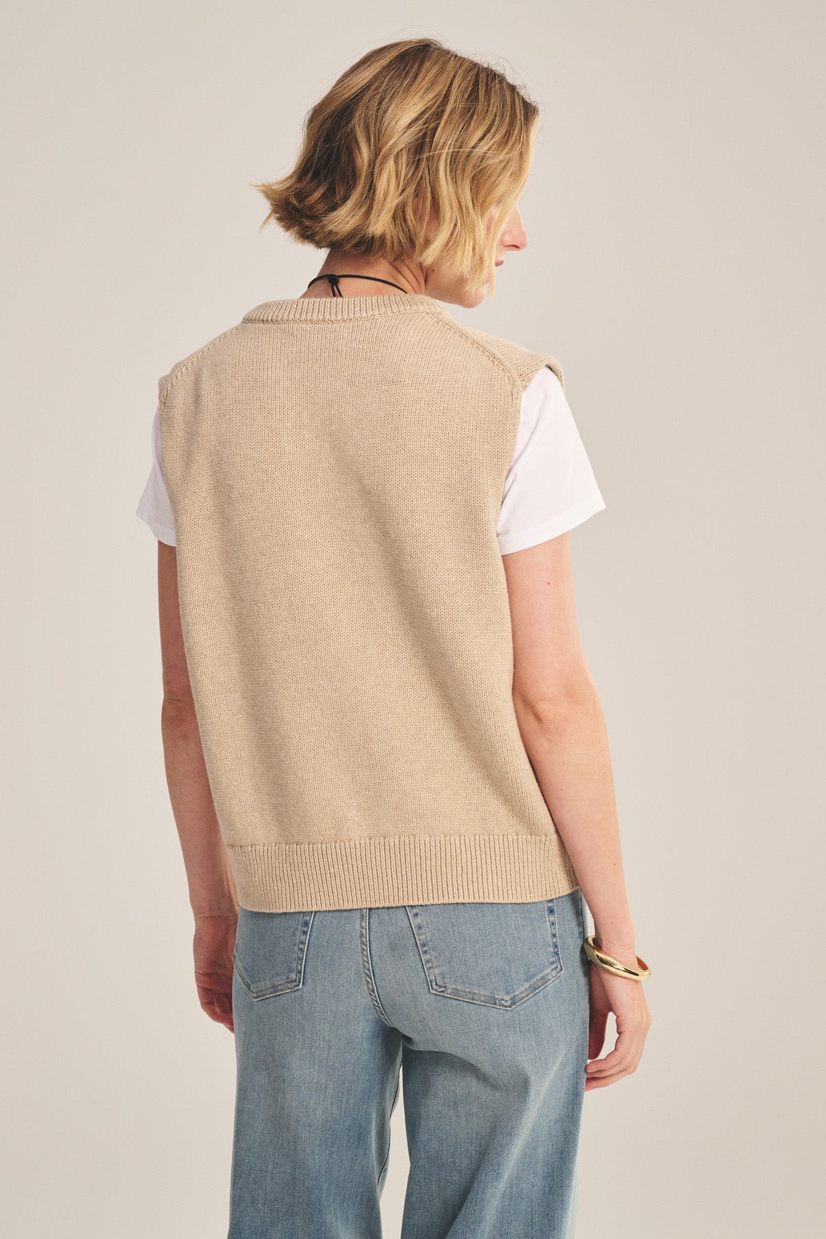 A person with short blonde hair, wearing the LUMINA SWEATER VEST by Velvet by Graham & Spencer over a white T-shirt and blue jeans, stands facing away from the camera, effortlessly showcasing a stylish layered outfit.