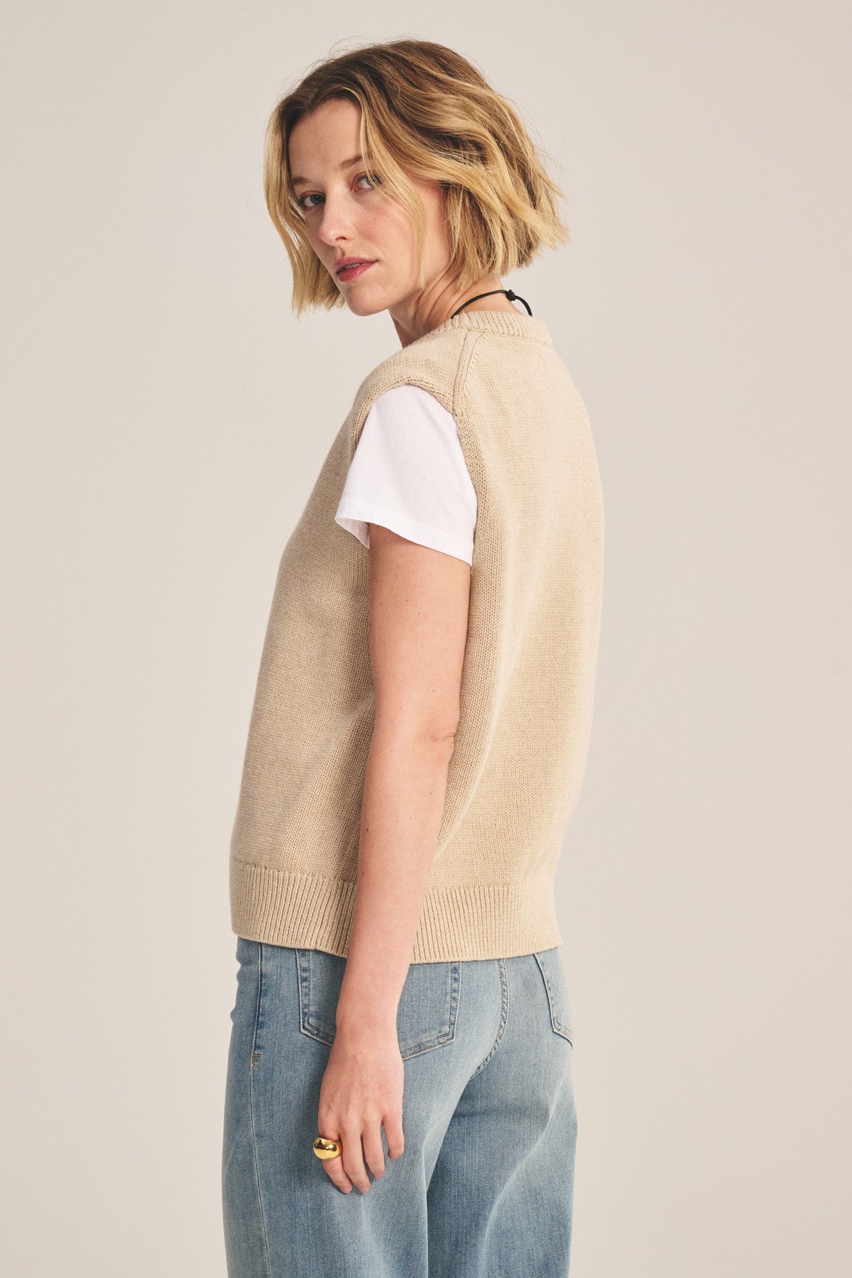 Wearing the LUMINA SWEATER VEST by Velvet by Graham & Spencer over a white tee and blue jeans, a person with short blonde hair stands sideways against a plain background, showcasing a stylish layered look.