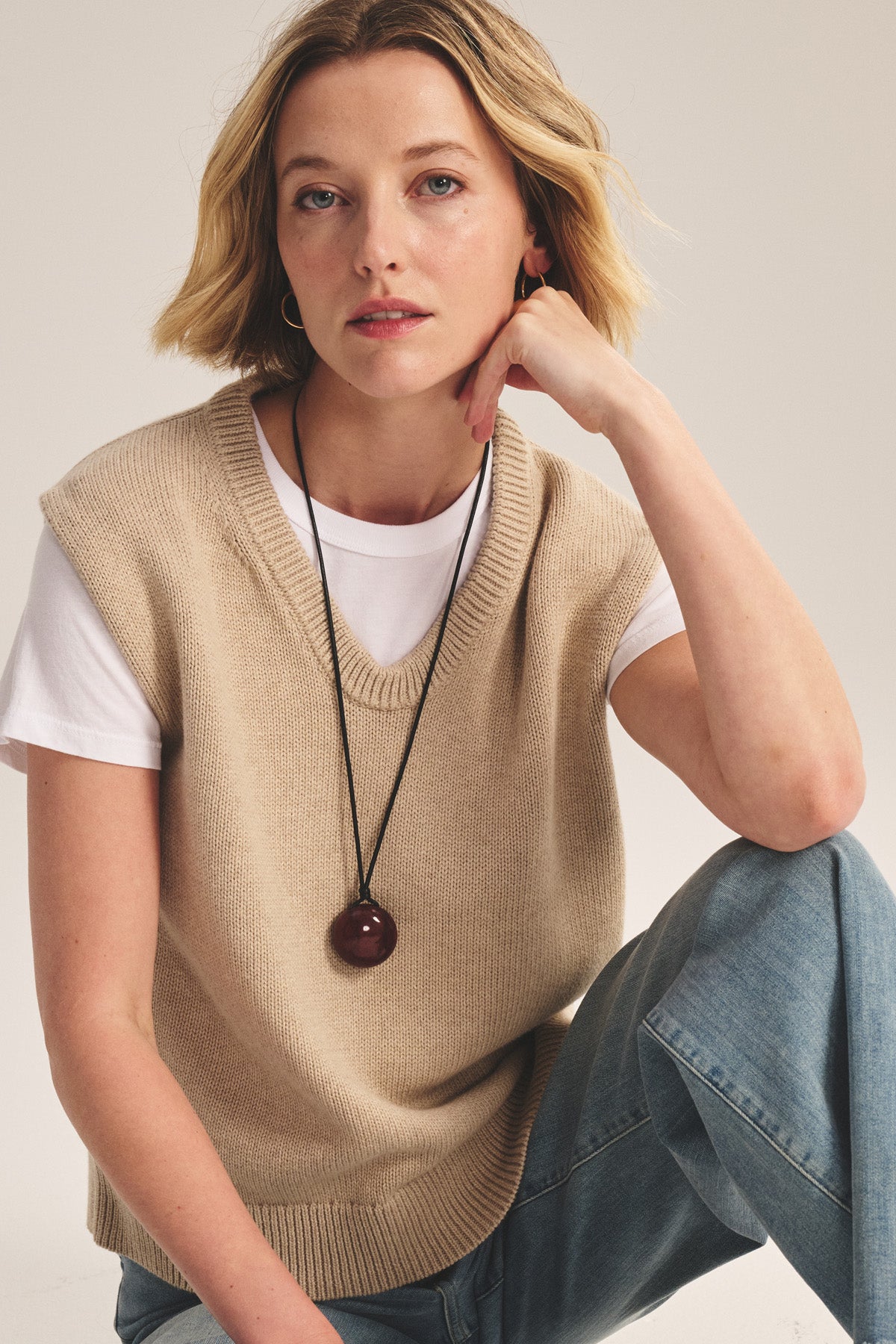 A woman with short blonde hair wears the LUMINA SWEATER VEST by Velvet by Graham & Spencer over a white tee, paired with blue jeans and a long dark pendant necklace, sitting casually with one knee up for an effortless layered look.-40909855752385