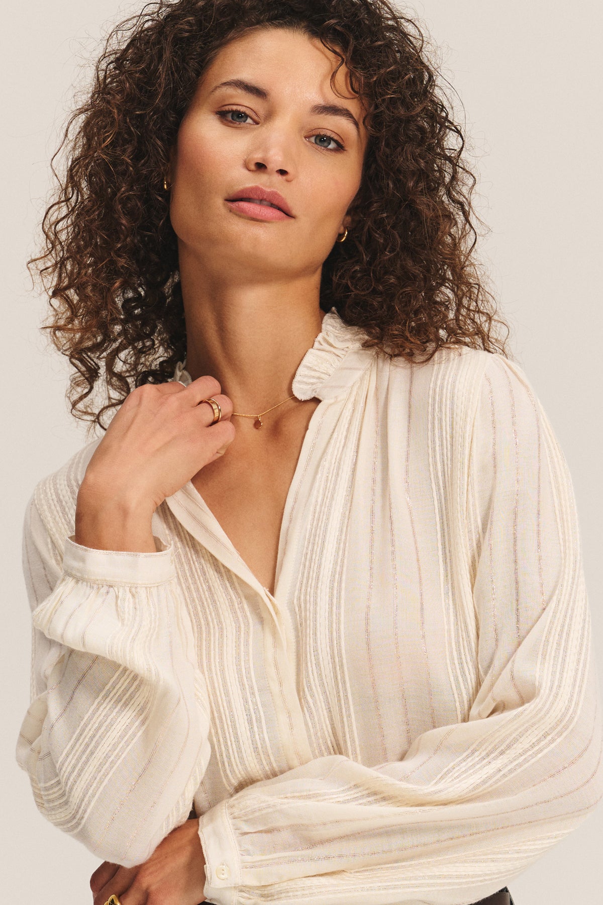 A woman with curly hair wears the cream CYNTHIA TOP by Velvet by Graham & Spencer, featuring ruffle details, looking at the camera with one hand touching her collar.-40247420354753