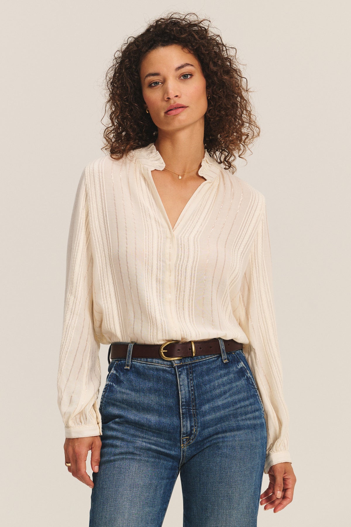 A woman with curly hair wears the cream CYNTHIA TOP by Velvet by Graham & Spencer, styled with high-waisted blue jeans and a dark brown belt, standing against a neutral background.
