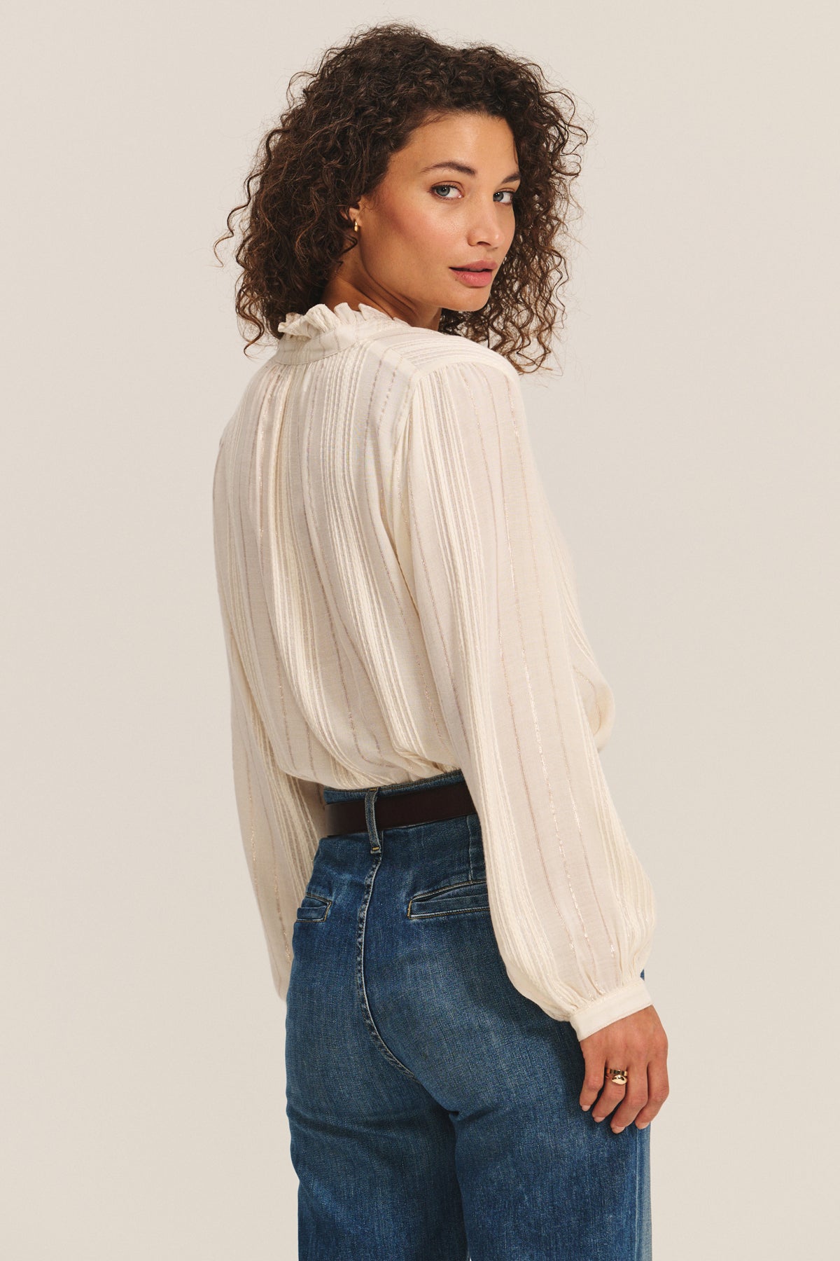 A woman with curly hair wears the CYNTHIA TOP by Velvet by Graham & Spencer—a cream long-sleeve blouse with ruffle detailing—paired with blue jeans, standing and looking over her shoulder against a neutral background.
