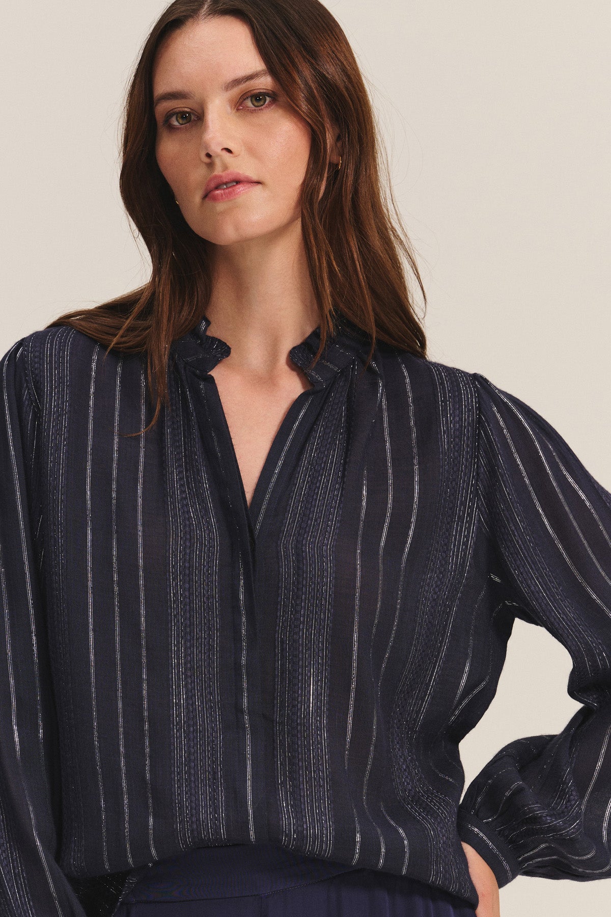 A woman with long brown hair wears the CYNTHIA TOP by Velvet by Graham & Spencer—a dark, long-sleeved viscose top with pinstripes, a slight V-neck, and gathered cuffs—standing against a light neutral background.