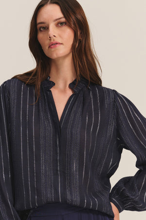 A woman with long brown hair wears the CYNTHIA TOP by Velvet by Graham & Spencer—a dark, long-sleeved viscose top with pinstripes, a slight V-neck, and gathered cuffs—standing against a light neutral background.