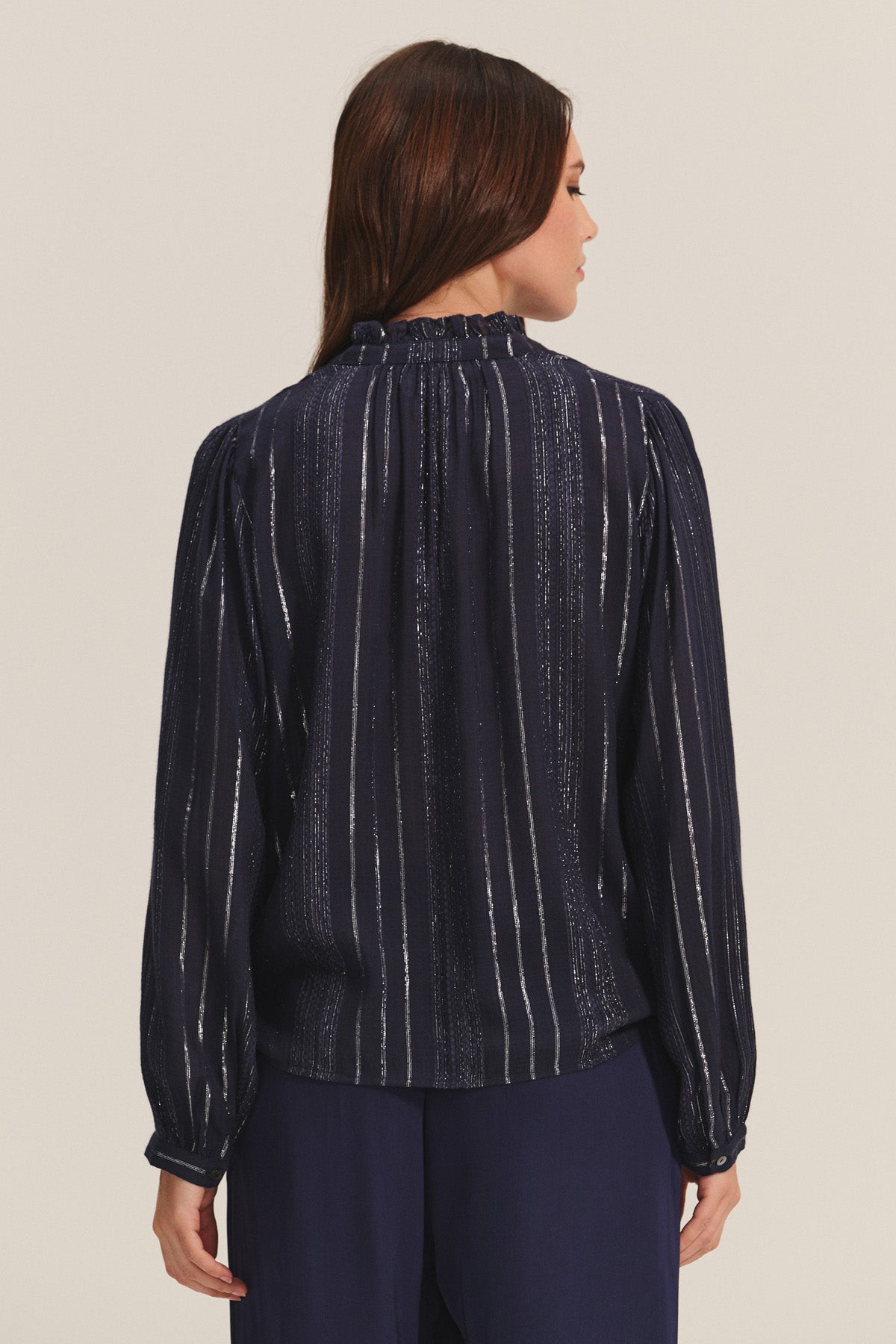 A woman is shown from the back wearing the CYNTHIA TOP by Velvet by Graham & Spencer—a navy, long-sleeved viscose top with vertical metallic lurex weaving—paired with loose navy pants against a neutral background.