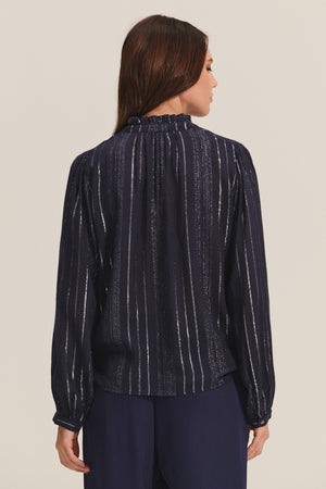 A woman is shown from the back wearing the CYNTHIA TOP by Velvet by Graham & Spencer—a navy, long-sleeved viscose top with vertical metallic lurex weaving—paired with loose navy pants against a neutral background.