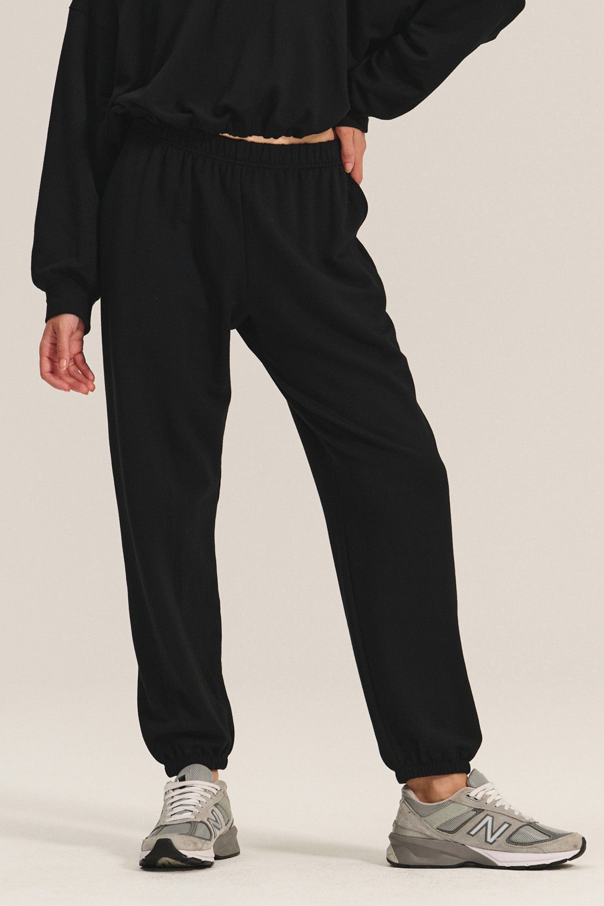 A person wears the EVERS JOGGER by Velvet by Graham & Spencer—relaxed fit black sweatpants with an elastic waist—paired with a black long-sleeve top and gray New Balance sneakers, standing against a plain background.-40334207090881