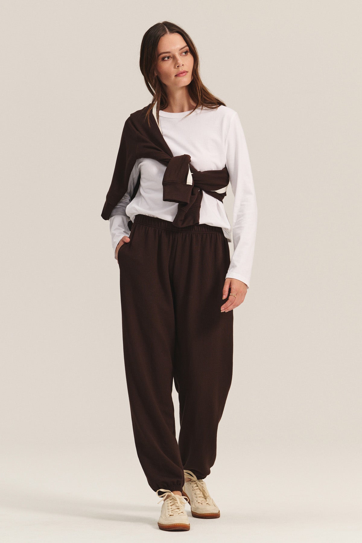 A woman wears the EVERS JOGGER by Velvet by Graham & Spencer—relaxed fit brown pants with an elastic waist—paired with a white long-sleeve shirt, a brown sweatshirt draped on her shoulders, and beige sneakers against a plain background.